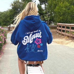 May include: A blue hoodie with a white graphic print that says "i left my heart in Maui" with a flower design and two people surfing. The text "A hui hou" is printed below the graphic.