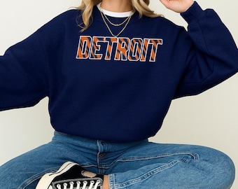May include: Navy blue sweatshirt with the word "DETROIT" in orange and white block letters. The sweatshirt is worn with blue jeans and black sneakers.