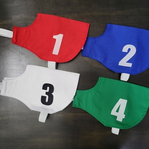 May include: Four colorful racing bibs in red, blue, white, and green, each with a large white number. The bibs have a curved shape and a white strap. Numbers are 1, 2, 3, and 4. The background is a dark wood surface.