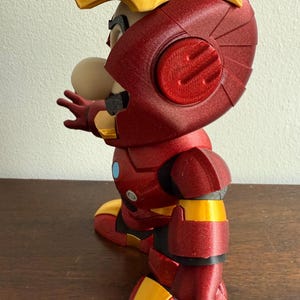 Peut inclure: Une figurine imprim&eacute;e en 3D rouge et or repr&eacute;sentant un personnage portant une armure d'Iron Man. La figurine est pos&eacute;e avec un bras tendu et l'autre bras pli&eacute; au coude.
