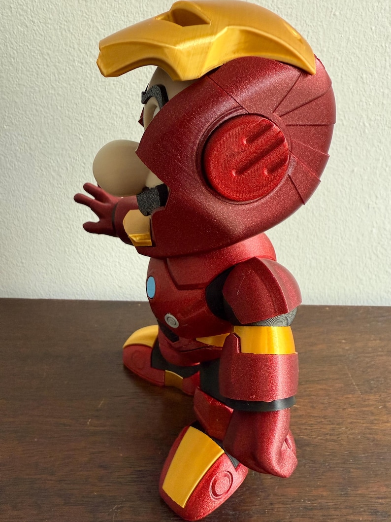 Peut inclure: Une figurine imprim&eacute;e en 3D rouge et or repr&eacute;sentant un personnage portant une armure d'Iron Man. La figurine est pos&eacute;e avec un bras tendu et l'autre bras pli&eacute; au coude.