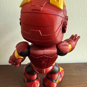 Peut inclure: figurine Iron Man rouge et or avec une surface textur&eacute;e. La figurine est debout, les bras tendus, et fait face &agrave; l'arri&egrave;re de la cam&eacute;ra.