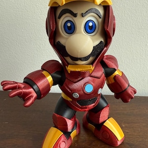 Peut inclure: Une figurine imprim&eacute;e en 3D rouge et or repr&eacute;sentant un personnage portant une armure rouge et or. Le personnage a une moustache et un grand nez. La figurine est debout sur une surface en bois.