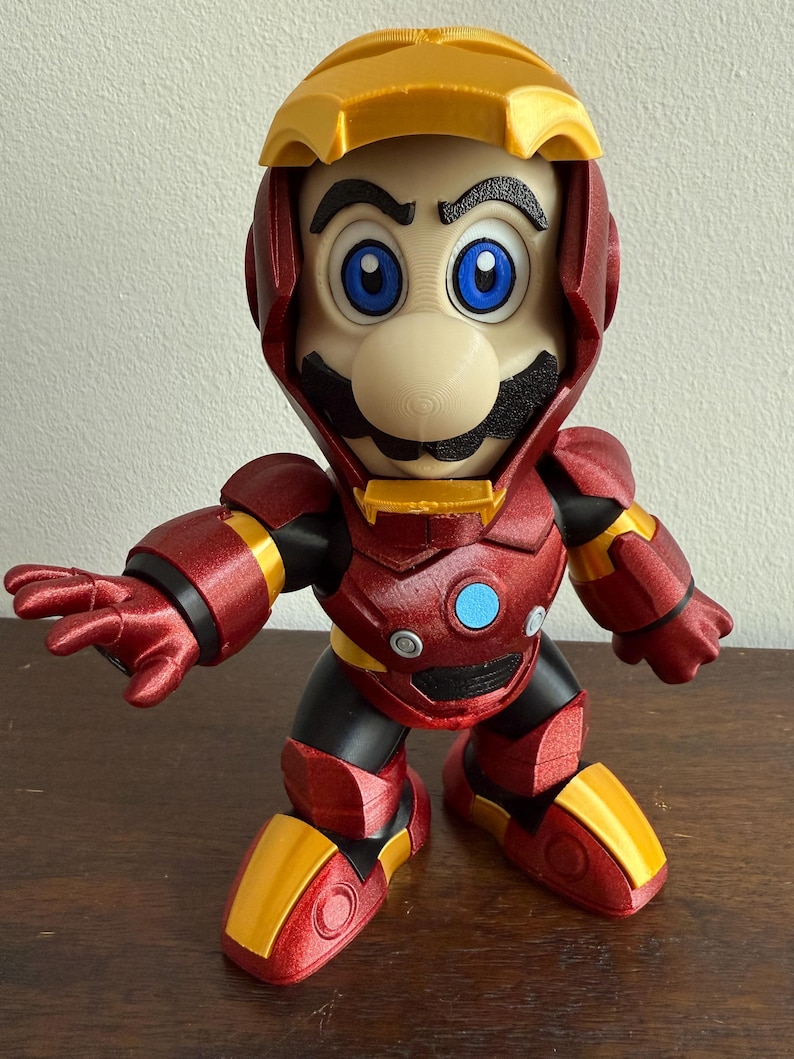 Peut inclure: Une figurine imprim&eacute;e en 3D rouge et or repr&eacute;sentant un personnage portant une armure rouge et or. Le personnage a une moustache et un grand nez. La figurine est debout sur une surface en bois.