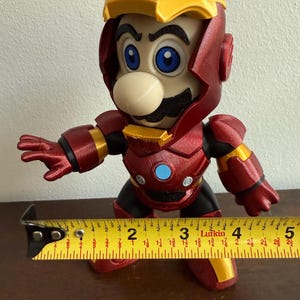Peut inclure: Une figurine rouge et or d'un personnage portant une armure d'Iron Man. Le personnage a une moustache et un grand nez. La figurine mesure environ 18 cm de haut.