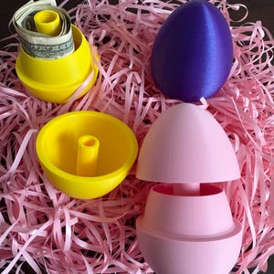 May include: Four plastic Easter eggs, two yellow, one pink, and one purple. The yellow eggs are open and one contains money. The pink egg is open and has a compartment inside. The eggs are sitting on a bed of pink shredded paper.