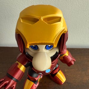 Peut inclure: Une figurine imprim&eacute;e en 3D d'un personnage portant une armure Iron Man rouge et or. Le personnage a un grand nez et des yeux bleus.