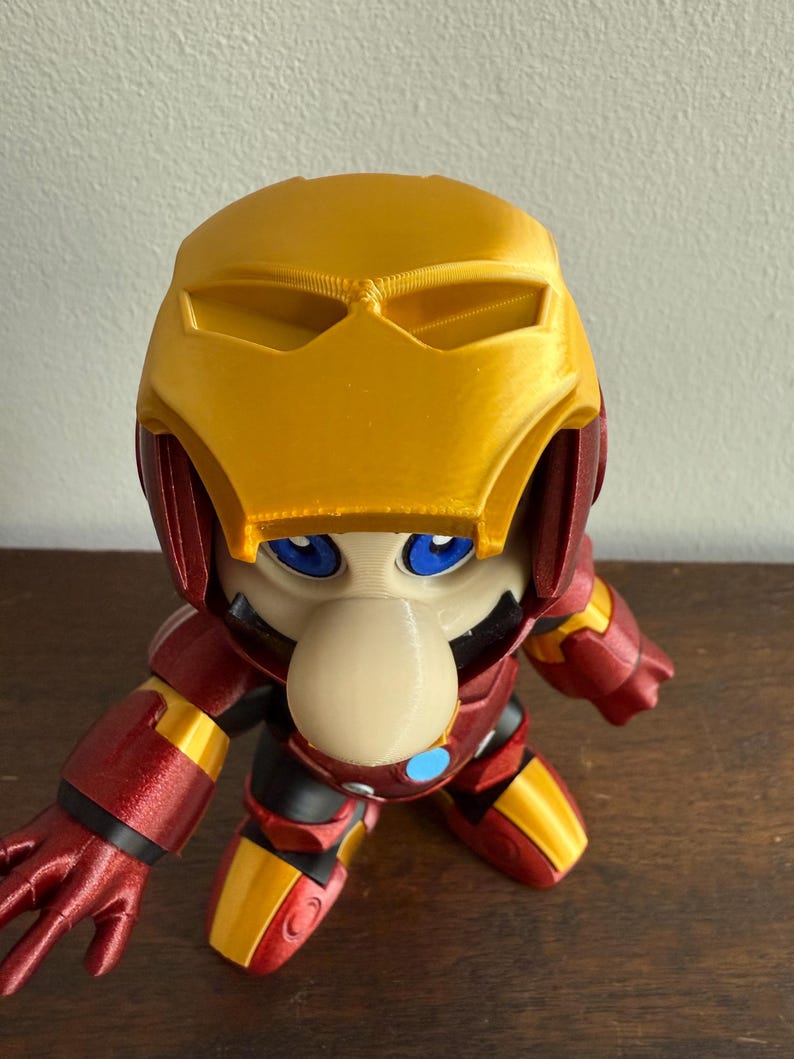 Peut inclure: Une figurine imprim&eacute;e en 3D d'un personnage portant une armure Iron Man rouge et or. Le personnage a un grand nez et des yeux bleus.