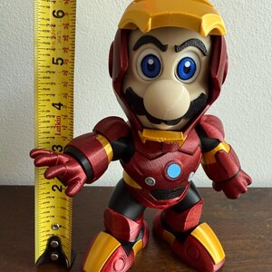 Peut inclure: Une figurine imprim&eacute;e en 3D rouge et or d'un personnage de jeu vid&eacute;o portant une armure Iron Man rouge et or. Le personnage a une moustache noire et un visage bleu et blanc.