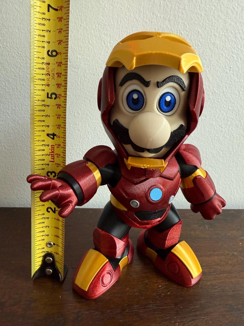 Peut inclure: Une figurine imprim&eacute;e en 3D rouge et or d'un personnage de jeu vid&eacute;o portant une armure Iron Man rouge et or. Le personnage a une moustache noire et un visage bleu et blanc.