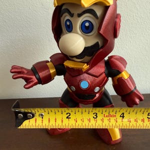 Peut inclure: Une figurine imprim&eacute;e en 3D rouge et or d'un personnage portant une armure d'Iron Man. Le personnage a une moustache et un grand nez. La figurine mesure environ 18 cm de haut.