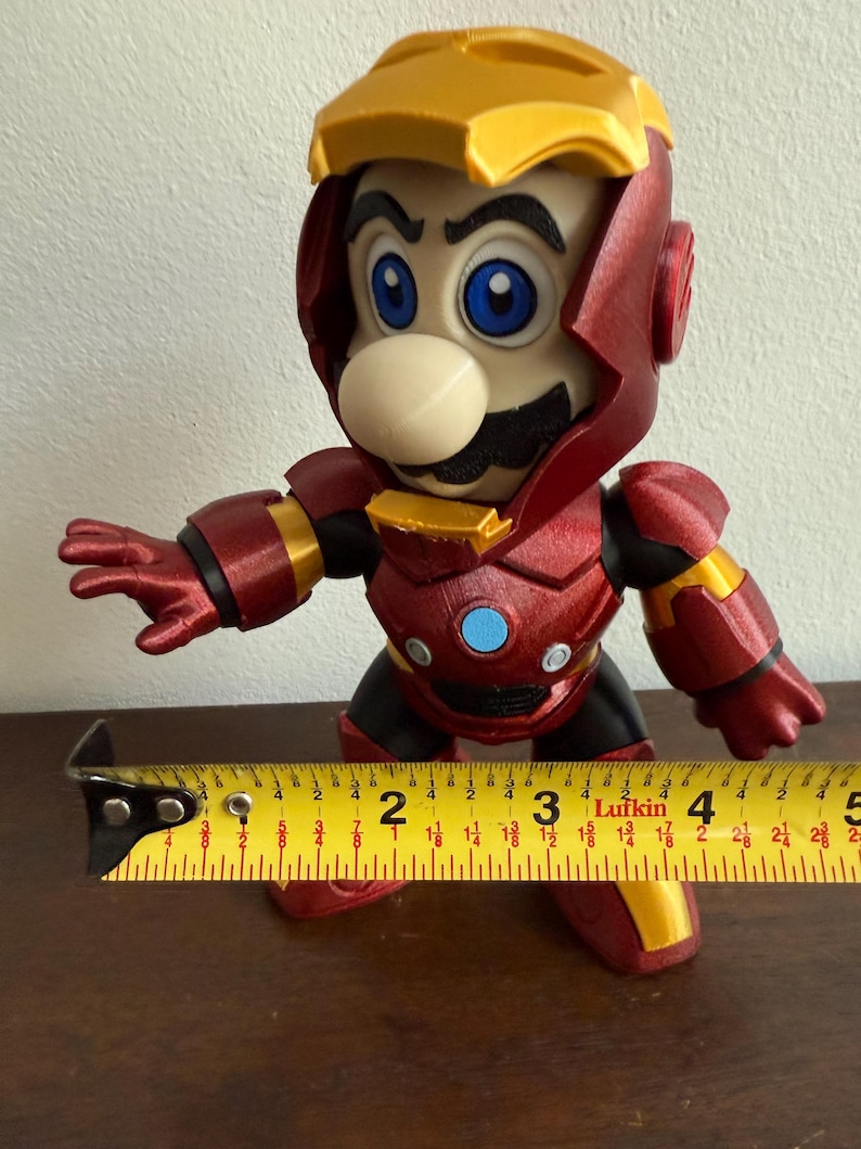 Peut inclure: Une figurine imprim&eacute;e en 3D rouge et or d'un personnage portant une armure d'Iron Man. Le personnage a une moustache et un grand nez. La figurine mesure environ 18 cm de haut.