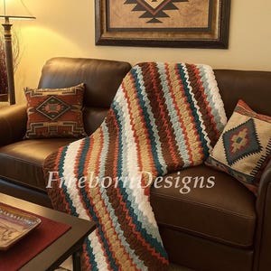 May include: A brown leather sofa with a colorful, striped crochet blanket draped over it. The blanket features stripes of white, teal, brown, and orange. Two decorative pillows with geometric patterns are on the sofa. The text "Fireborn Designs" is on the blanket.