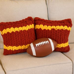 May include: Two red and yellow striped decorative pillows and a brown football with white stripes are displayed on a beige couch. The pillows have a chunky knit texture, and the football is positioned in front of them.