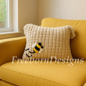 May include: A cream-colored, textured pillow with a crocheted bee applique, resting on a yellow sofa. The bee has black and yellow stripes. The pillow has a square shape with small tassels on the corners. The text "Freeborn Designs" is visible.