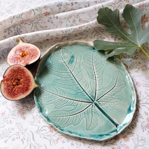 Op de afbeelding: Een turquoise keramische serveerschaal met een reliëfdesign van een vijgenblad. Twee rijpe vijgen liggen op een witte, bloemige tafelkleed op de achtergrond.