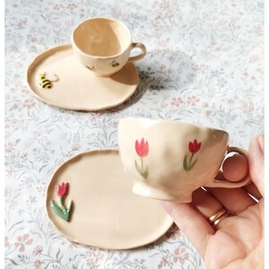 Può includere: Due tazze da tè in ceramica e piattini con disegni floreali e di api. Le tazze da tè sono di colore crema con tulipani rossi e api dipinte su di esse. I piattini sono anche di colore crema e hanno un'ape e un tulipano dipinti su di essi.