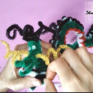 Puede incluir: Un drag&oacute;n de limpiapipas verde, negro, blanco y rojo con ojos saltones. El drag&oacute;n est&aacute; siendo sostenido por las manos de una persona. La cola del drag&oacute;n est&aacute; siendo cortada con alicates. El texto "Art Studio Shevchenko" es visible en la esquina superior derecha de la imagen.