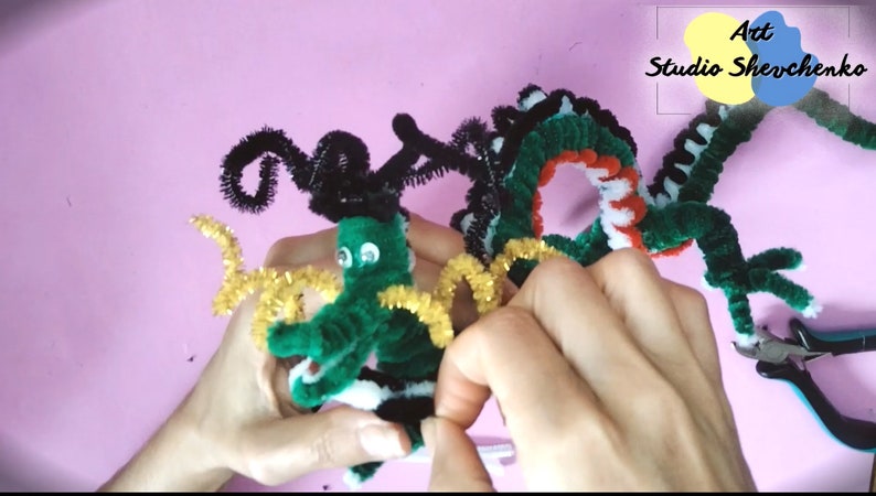 Puede incluir: Un drag&oacute;n de limpiapipas verde, negro, blanco y rojo con ojos saltones. El drag&oacute;n est&aacute; siendo sostenido por las manos de una persona. La cola del drag&oacute;n est&aacute; siendo cortada con alicates. El texto "Art Studio Shevchenko" es visible en la esquina superior derecha de la imagen.