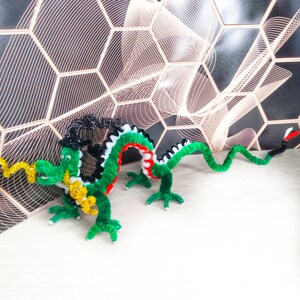 Puede incluir: Un drag&oacute;n de limpiapipas verde, negro y blanco con una cola de limpiapipas amarilla y dorada. El drag&oacute;n est&aacute; en posici&oacute;n con la cabeza girada hacia un lado y la cola enroscada detr&aacute;s de &eacute;l.