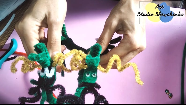 Puede incluir: Dos ara&ntilde;as de limpiapipas verdes con patas negras y ojos saltones. Las ara&ntilde;as est&aacute;n conectadas por un limpiapipas amarillo. El texto "Art Studio Shevchenko" es visible en el fondo.