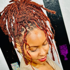 May include: A woman with reddish-brown locs styled up. The hair is a vibrant shade of auburn with lighter red highlights. She is wearing a beige sweater and gold jewelry. The background has black and white stripes.