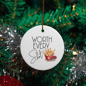 May include: A white, round Christmas ornament with the text "Worth Every Shot" and a graphic of fries and ketchup. The ornament is hanging on a Christmas tree with red and silver decorations.
