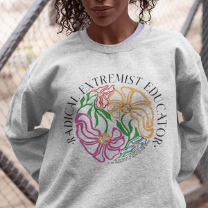 May include: A grey sweatshirt with a circular graphic design featuring stylized flowers and the text "Radical Extremist Educator".