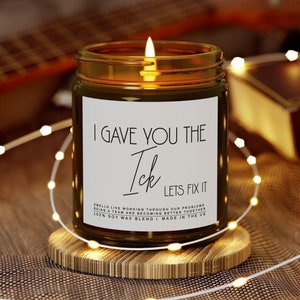 Puede incluir: Una vela encendida en un tarro de cristal con una etiqueta blanca que dice "I GAVE YOU THE Ick LETS FIX IT". La vela está sobre un posavasos de madera, rodeada de luces de cadena. El fondo incluye una guitarra y un libro.