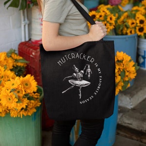 Op de afbeelding: Zwarte draagtas met een skeletballetdanseres en notenkraker illustratie, met de tekst "Nutcracker is my favorite season". De tas wordt vastgehouden door een persoon voor een bloemen display.