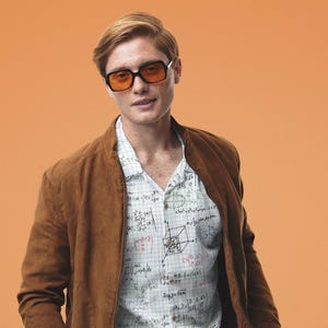 May include: A man wearing a brown suede jacket over a white button-down shirt with a pattern of mathematical equations and formulas. He is wearing orange-tinted sunglasses and brown trousers.