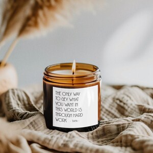 May include: A lit candle in a brown glass jar with a white label. The label reads, "THE ONLY WAY TO GET WHAT YOU WANT IN THIS WORLD IS THROUGH HARD WORK - TIANA." The candle sits on a beige cloth.