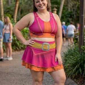 Puede incluir: Un conjunto deportivo de dos piezas que incluye un sujetador deportivo rosa y naranja y una falda a juego con un diseño de cinturón estampado. El conjunto se combina con zapatillas deportivas rosas y naranjas y una diadema con dos orejas redondas rosas y un lazo.