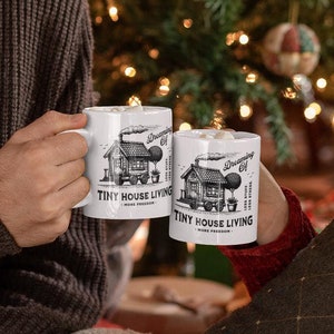 May include: Two white ceramic mugs with a black and white illustration of a tiny house and the text "TINY HOUSE LIVING" are held up in a toast. The mugs are held by two people in front of a Christmas tree.