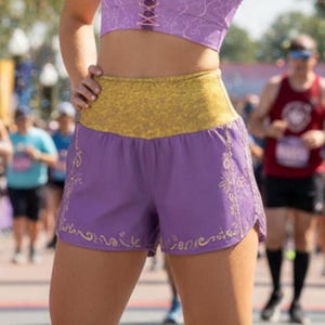 Puede incluir: Un top deportivo corto de color morado con cordones en la parte delantera y un sutil patrón de sol, combinado con pantalones cortos deportivos morados a juego con una cintura dorada texturizada y bordados florales dorados en los dobladillos.
