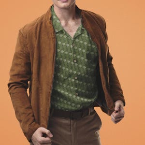 May include: A brown suede bomber jacket with a zipper closure, worn over a green button-down shirt with a white floral pattern. The outfit includes brown pants and a brown belt. The background is a solid orange.