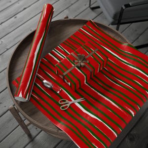 May include: Red wrapping paper with green and white brushstroke stripes. A wrapped gift with a brown ribbon sits on the paper, along with a roll of paper, scissors, and tape. The items are on a wooden table.