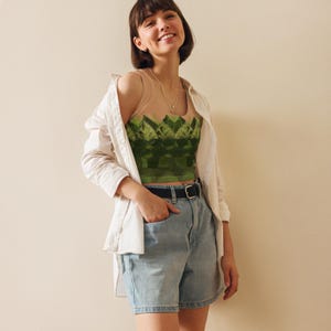 May include: A beige tank top with a green geometric design is worn with light wash denim shorts, a black belt, and a white button-down shirt. The tank top features a repeating pattern of green shapes. A gold necklace is also visible.