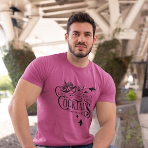 以下が含まれることがあります： 「Cupid's Cocktails & Love Potion」の黒い文字がプリントされた、マゼンタ色のクルーネックTシャツ。デザインにはキューピッドのイラストやその他の装飾要素が含まれています。画像内の人物はブルージーンズを着用しています。