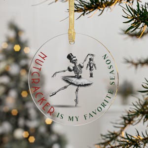 May include: Clear, round Christmas ornament with a gold ribbon. The ornament features a skeleton ballerina holding a nutcracker, with the text "Nutcracker is my favorite season" in red and green.