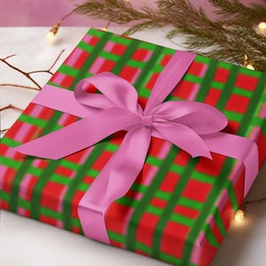 May include: A wrapped gift with a pink bow. The wrapping paper has a red, green, and pink plaid pattern. The bow is a solid pink color. The gift is on a white surface with a pink background.