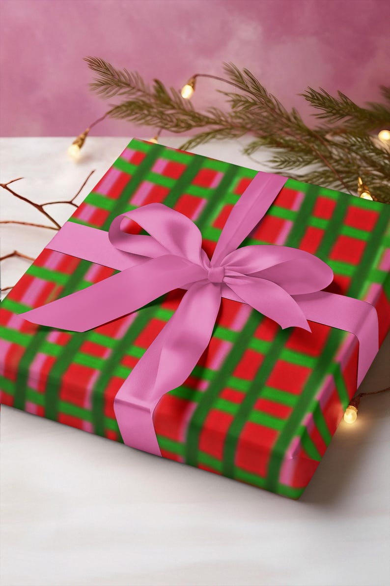 May include: A wrapped gift with a pink bow. The wrapping paper has a red, green, and pink plaid pattern. The bow is a solid pink color. The gift is on a white surface with a pink background.