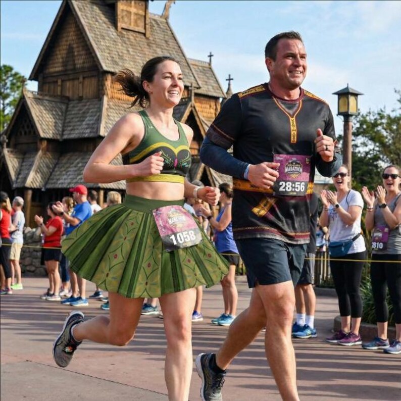 May include: A man and woman running a marathon. The woman wears a green and gold sports bra and a green skirt with a floral design. The man wears a black shirt with gold accents. Both have race bibs.