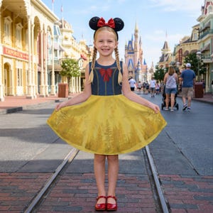 Puede incluir: Una niña con un vestido amarillo y azul con un lazo rojo y un estampado de bosque en la falda. También lleva una diadema negra con orejas de Minnie Mouse y zapatos rojos. El fondo muestra una calle con edificios y gente.