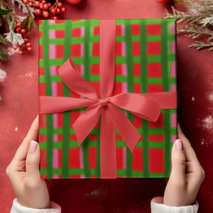 May include: A wrapped gift with a red ribbon and bow. The wrapping paper has a green and red plaid pattern. The gift is held by hands wearing a white sweater, set against a red background with Christmas decorations.