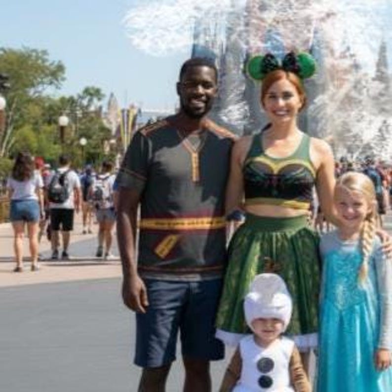 May include: A family poses outdoors, dressed in themed costumes. The adults wear green and gold outfits, while the children are dressed as characters. The background shows a sunny day with other people walking around.