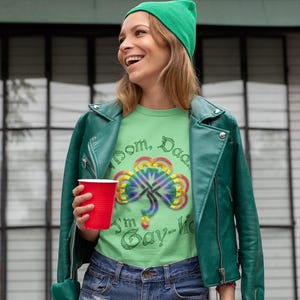 May include: A woman wearing a green leather jacket, a green shirt with a rainbow shamrock and the text "Mom, Dad, I'm Gay-lic", and denim shorts. She is holding a red cup.