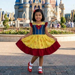 Puede incluir: Una niña con un vestido amarillo, azul y rojo, un lazo rojo y zapatos a juego. El vestido tiene una falda amarilla con estampado floral y un volante rojo en el dobladillo. Se ve un castillo al fondo.