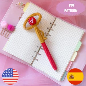 Puede incluir: Un bolígrafo de ganchillo hecho a mano con un diseño rojo y dorado, apoyado en un cuaderno abierto. El bolígrafo presenta un cuerpo rojo, un lazo dorado y una pequeña bola roja y blanca en la parte superior. Las palabras "PDF PATTERN" son visibles.