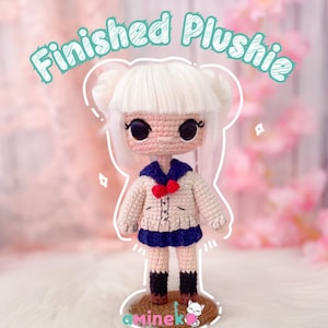 Può includere: Bambola all'uncinetto fatta a mano, con capelli bianchi e grandi occhi neri. La bambola indossa una giacca color crema con colletto e gonna blu e un fiocco rosso. Il testo "Finished Plushie" è sopra.